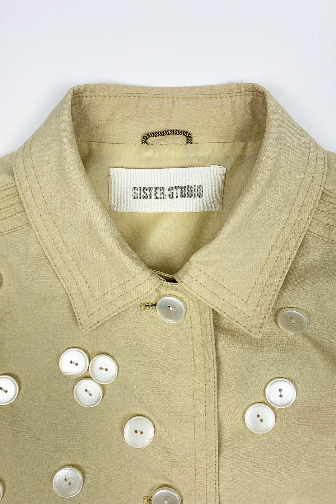 Close-up of a beige jacket with button details and 'Sister Studio' label on a white background