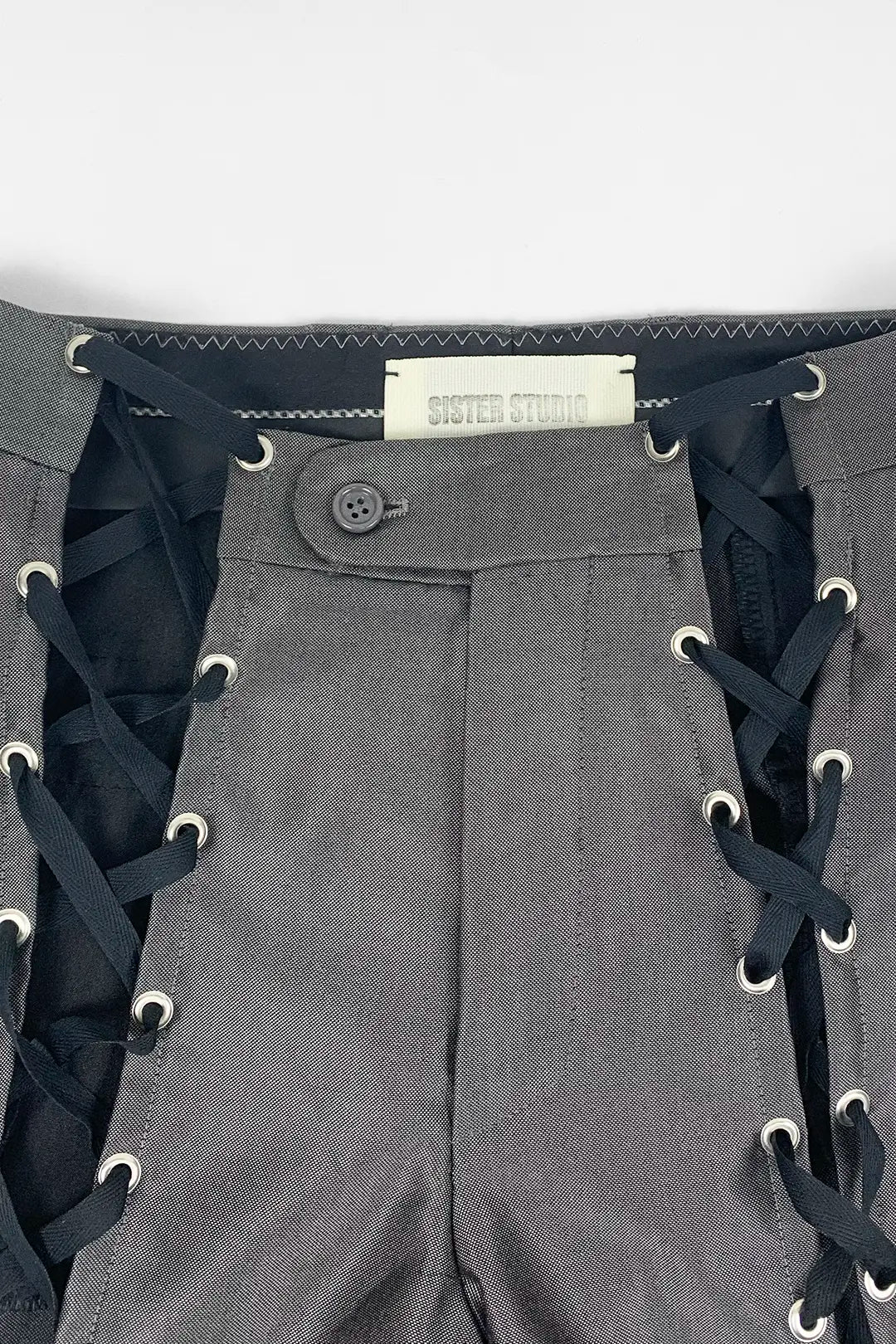 Close-up of gray pants with black lace-up side details, metal eyelets, a button closure, and a visible Sister Studio label.