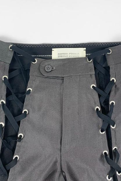 Close-up of gray pants with black lace-up side details, metal eyelets, a button closure, and a visible Sister Studio label.