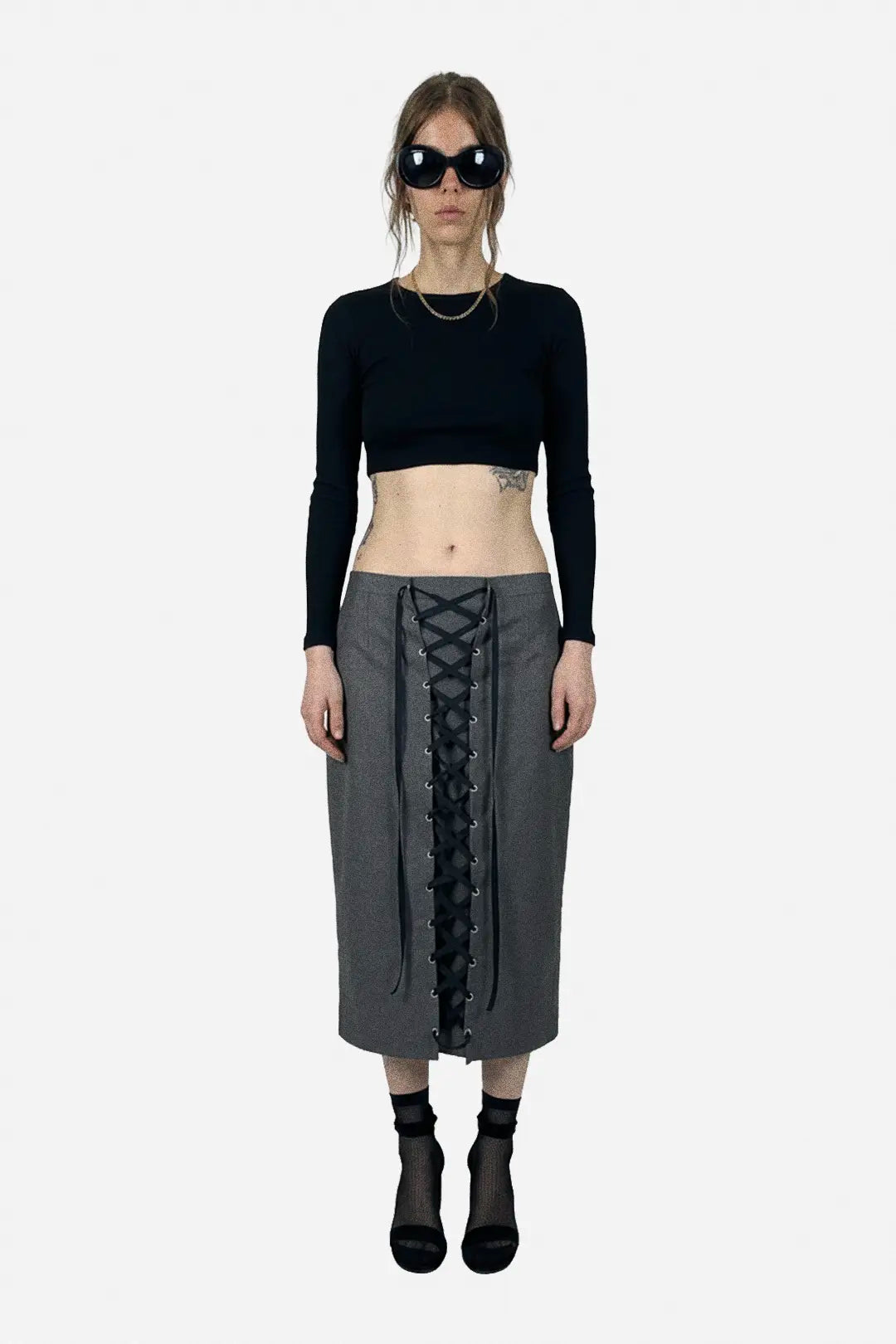 A model stands against a light gray background wearing a black long-sleeve crop top, a gray midi skirt with a black lace-up front, black socks, and black boots.
