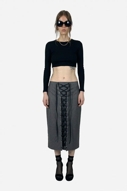 A model stands against a light gray background wearing a black long-sleeve crop top, a gray midi skirt with a black lace-up front, black socks, and black boots.