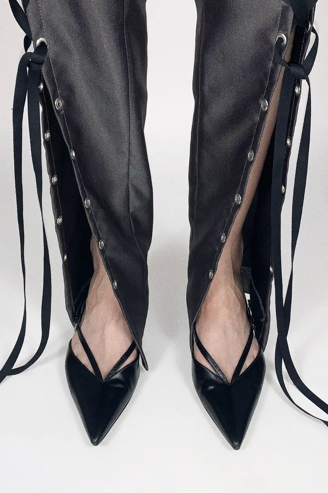 Black pointed-toe high heels with strappy side details and long ties, shown worn with matching black pants against a light gray studio background.