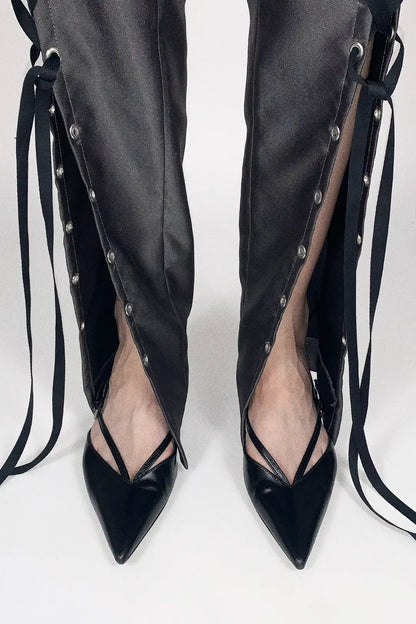 Black pointed-toe high heels with strappy side details and long ties, shown worn with matching black pants against a light gray studio background.