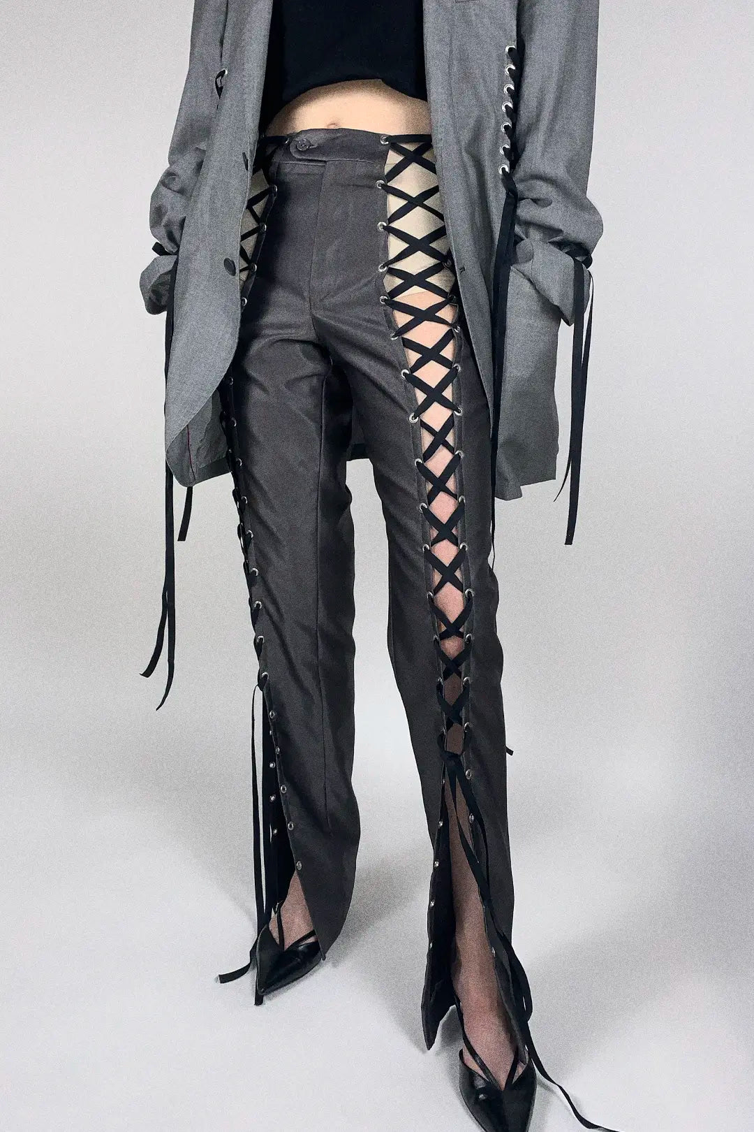 A model is shown from the waist down wearing dark gray high-waisted pants with prominent side criss-cross lace-up detailing and black pointed-toe heels, styled with a gray blazer.