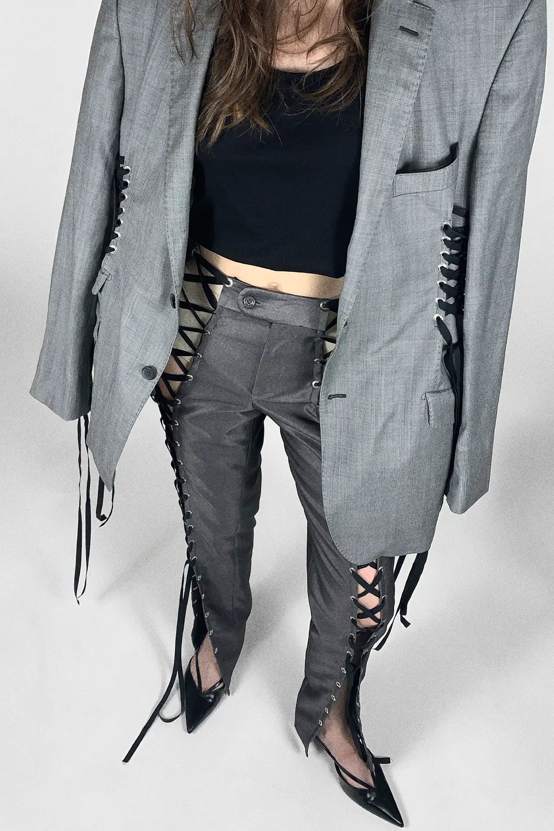 A model wearing a gray oversized blazer with black lace-up detailing down the sides, paired with matching gray lace-up pants and black pointed-toe heels, against a light gray studio background.