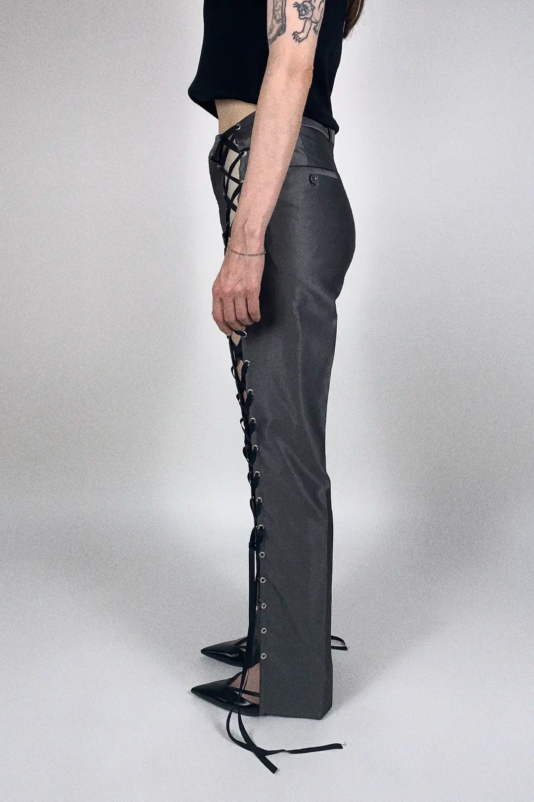 A side view of a model wearing dark gray high-waisted flared pants with black lace-up detailing running down the sides, paired with a black crop top and black pointed-toe heels, against a light gray studio background.