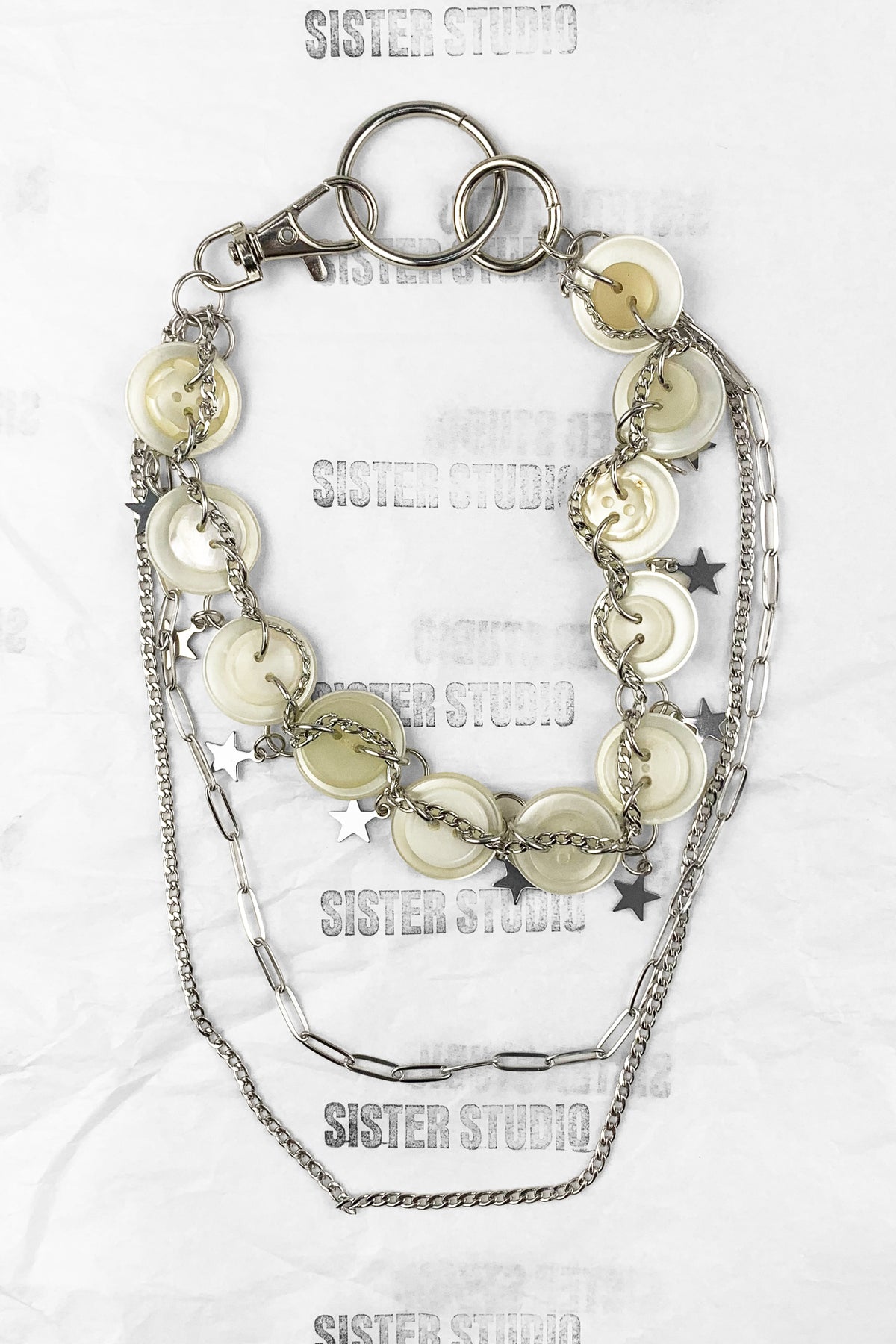 A layered silver-tone keychain necklace featuring round shell-like discs, star charms, paperclip chain, and a lobster clasp, displayed on a white textured background with 'SISTER STUDIO' watermarks.