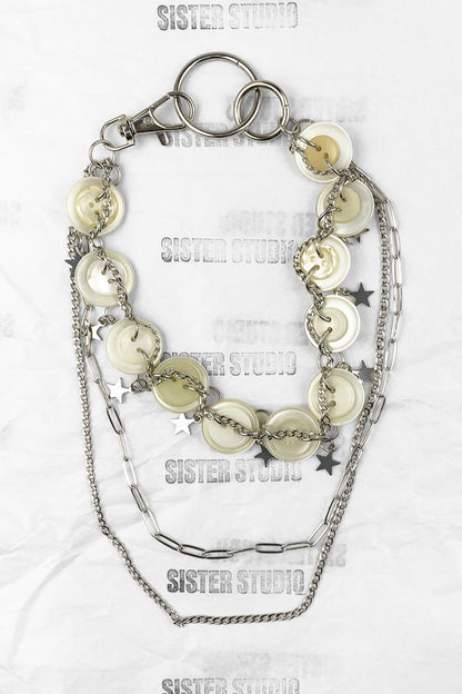 A layered silver-tone keychain necklace featuring round shell-like discs, star charms, paperclip chain, and a lobster clasp, displayed on a white textured background with 'SISTER STUDIO' watermarks.