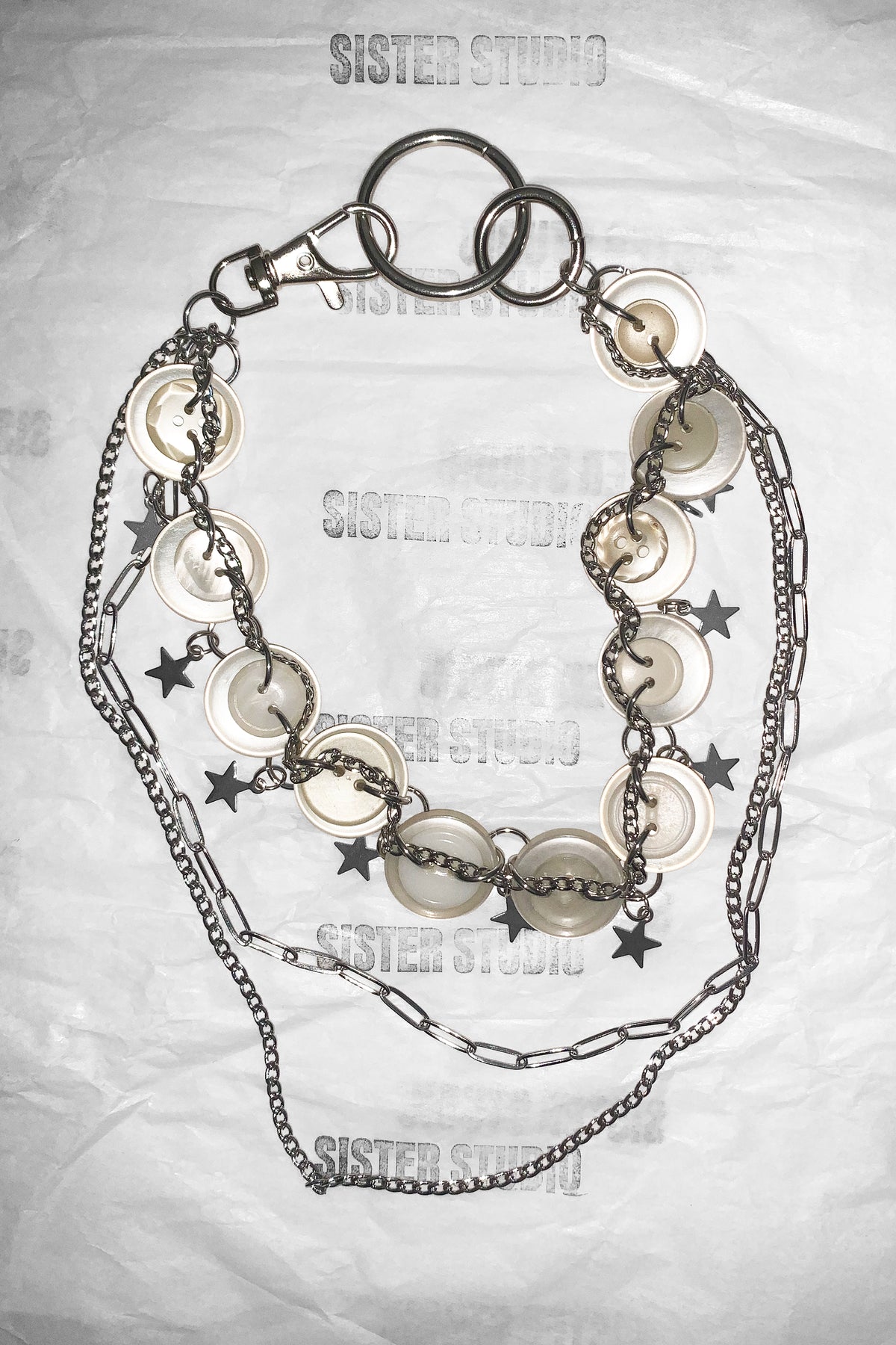 A layered chain necklace with silver-tone metal links, round white disc charms, and small black star charms, laid flat on a wrinkled white paper background with 'SISTER STUDIO' watermarks.