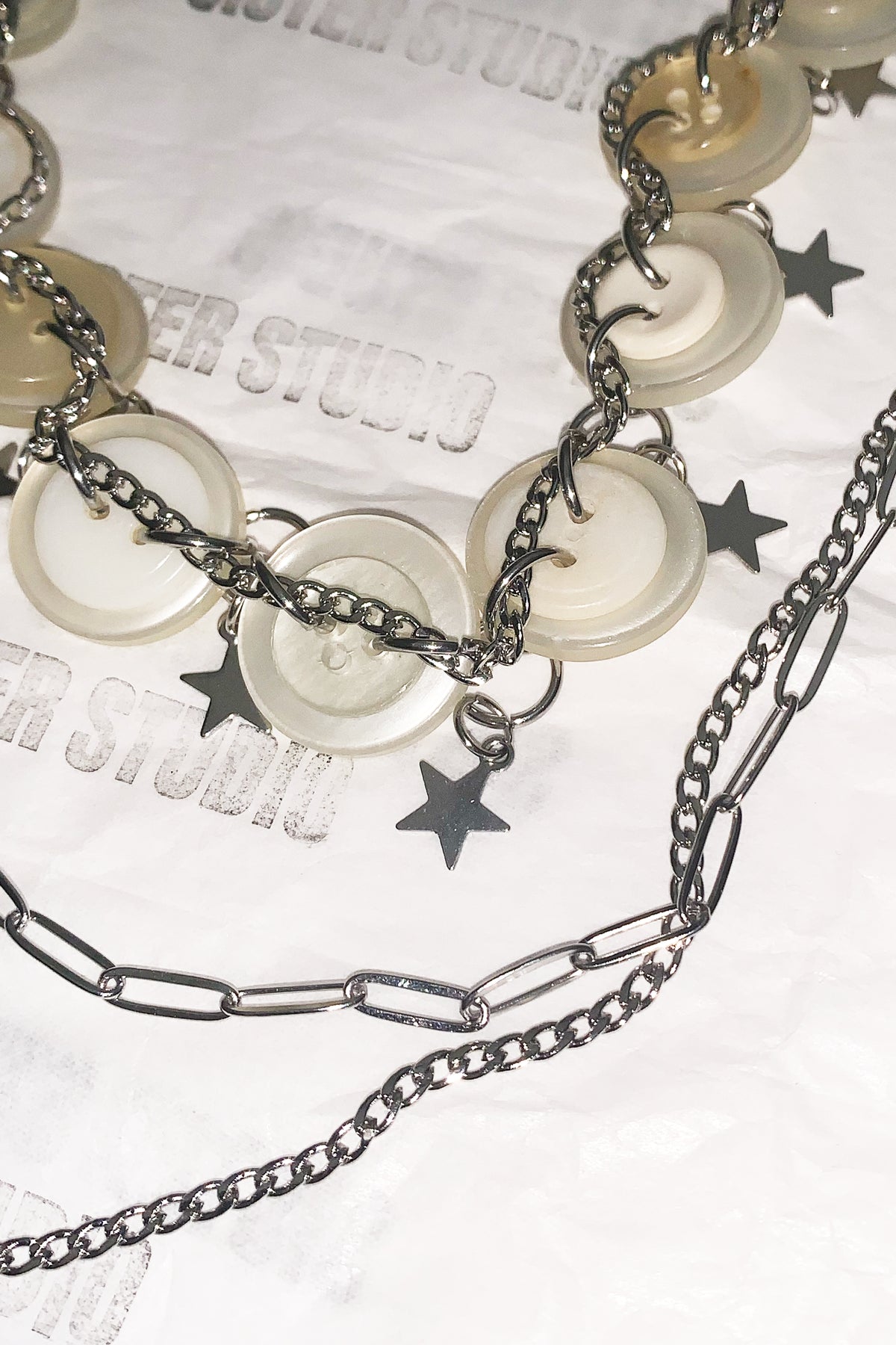 A close-up of a layered necklace featuring translucent round shell-like discs, dark metal chains, and small star charms, laid on white branded paper.