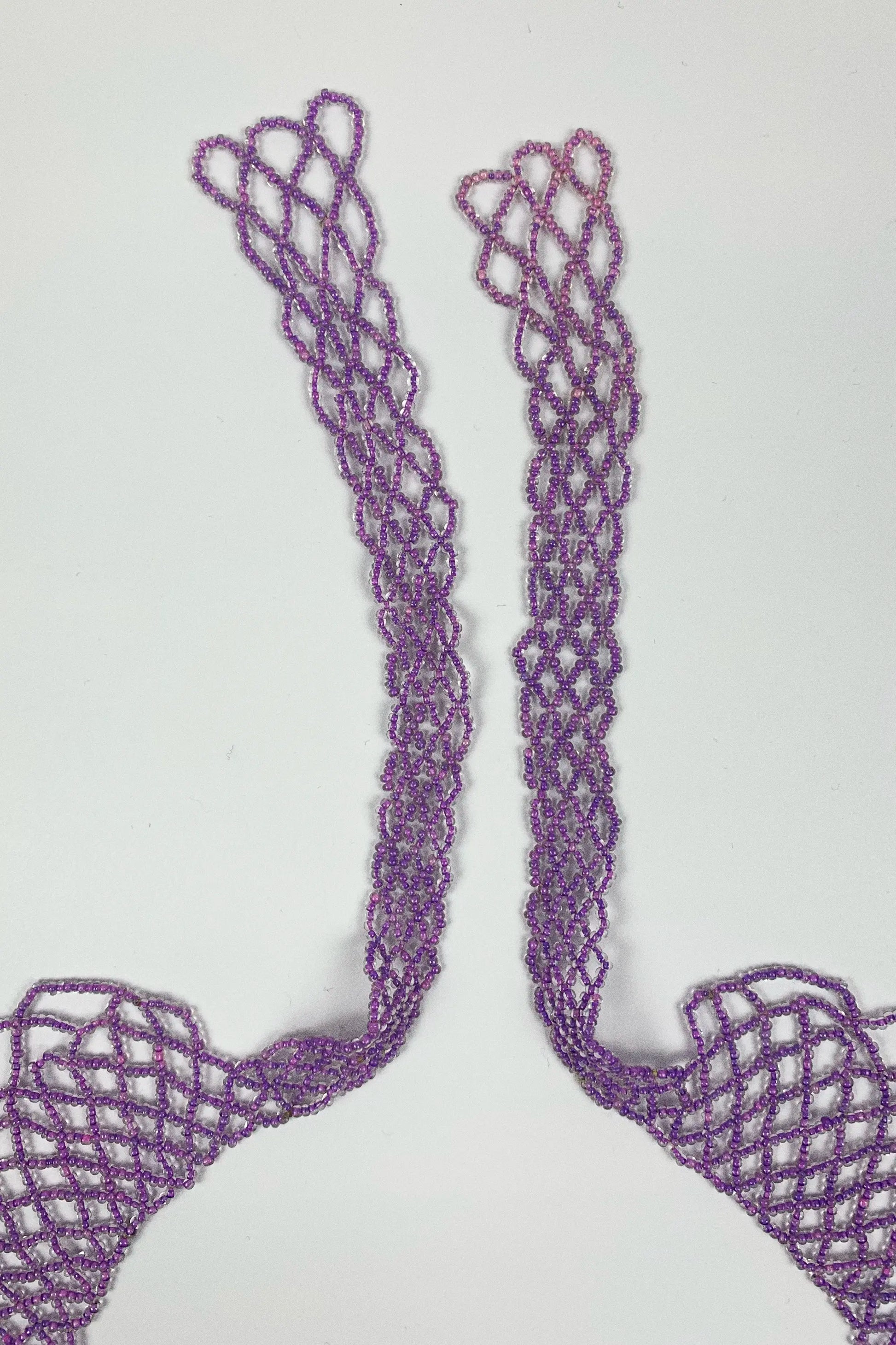 A pair of lavender purple crochet or lace trim pieces with a textured openwork pattern, laid flat on a light gray background.
