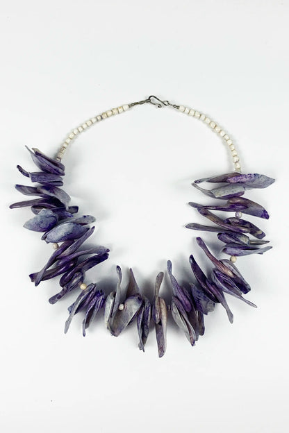 A statement necklace featuring irregular, jagged purple and gray stone-like pieces arranged in a circle, connected by a white beaded chain with a silver clasp, shown on a plain white background.