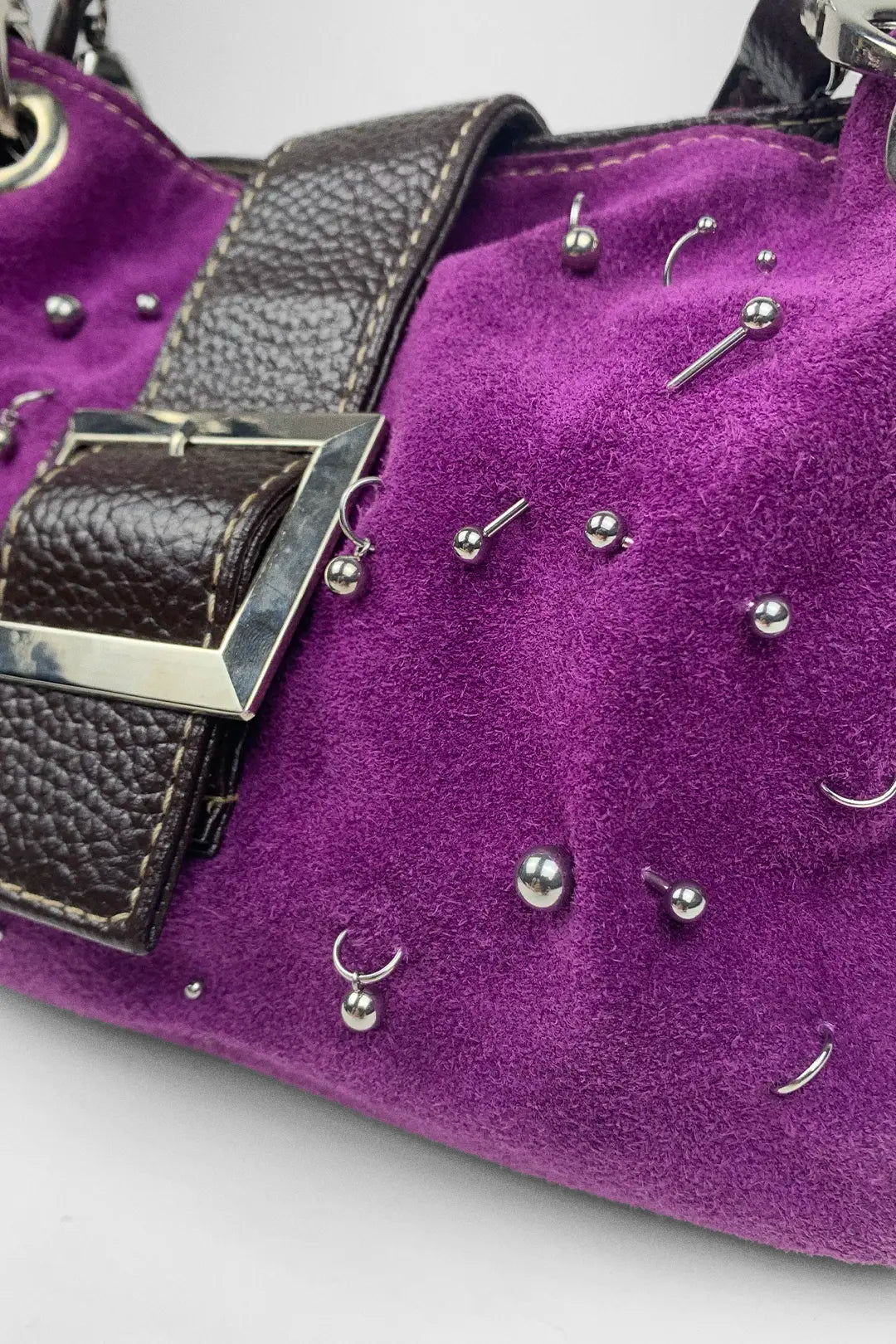 Close-up of a purple suede handbag with silver studs, crescent-shaped studs, and a dark brown textured strap with a silver buckle.