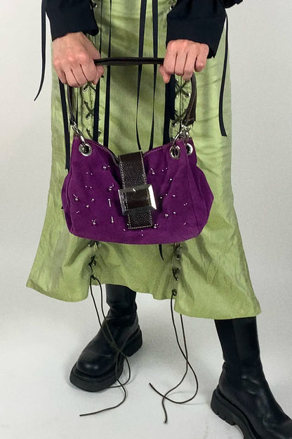 A person wearing a light green skirt with black lace-up details and black boots holds a purple suede shoulder bag with silver studs and a black leather strap detail against a light gray studio background.