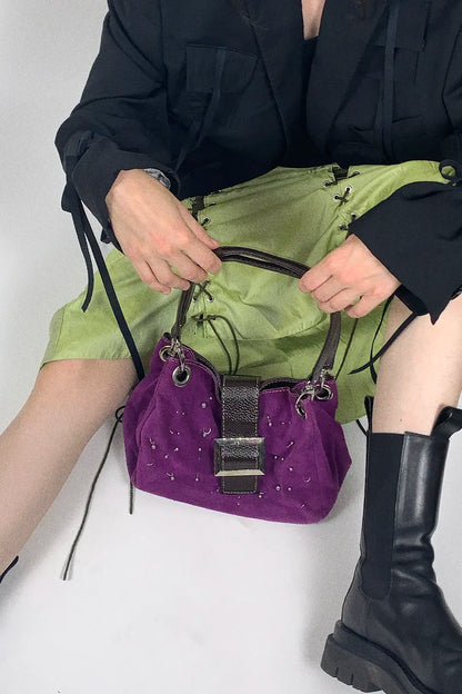 A person wearing a black jacket and lime green skirt holds a purple velvet handbag with silver studs and a dark metallic front clasp. The person is also wearing black leather boots against a plain white background.