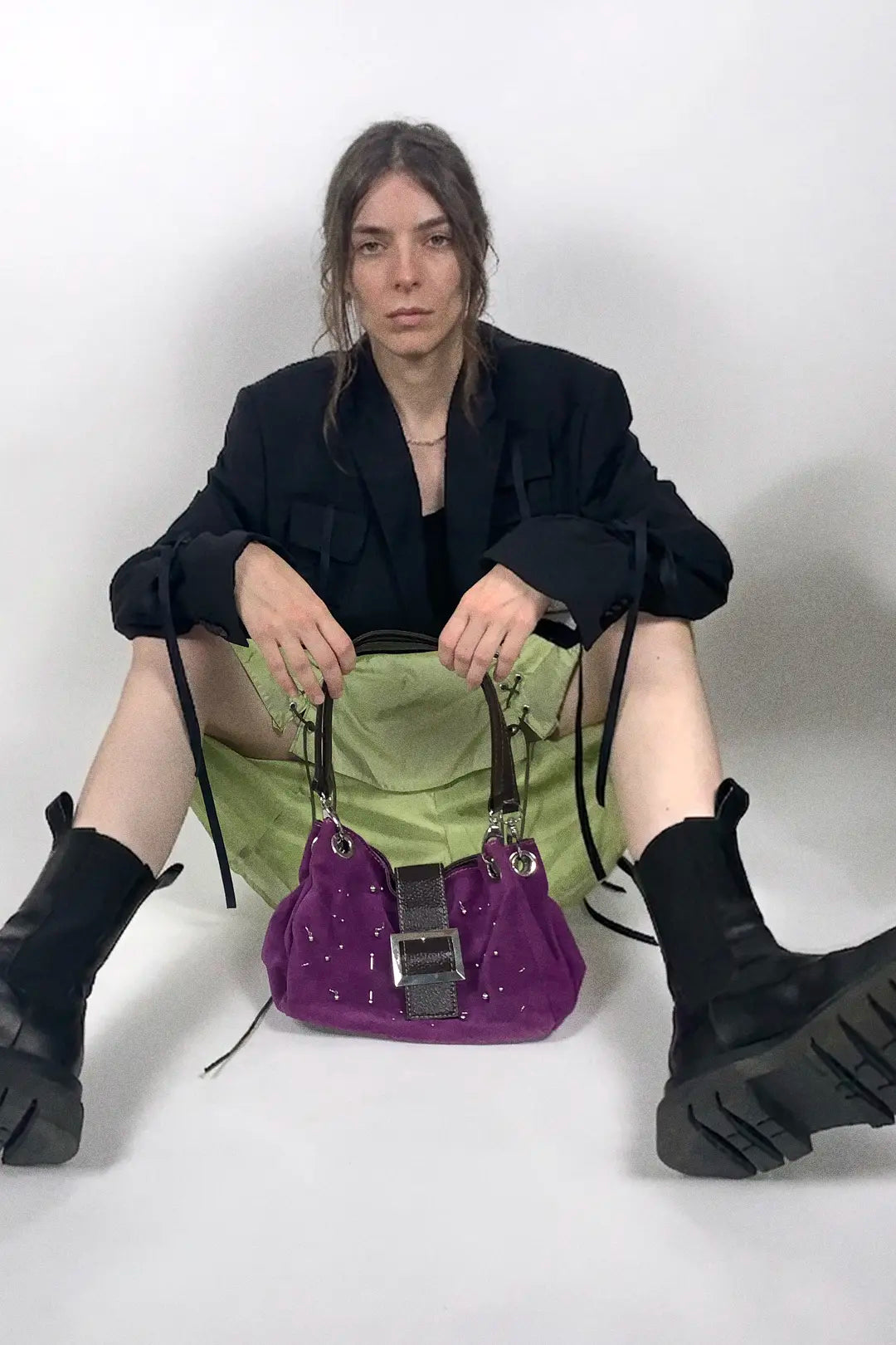 A model sits on a white floor against a white wall, wearing a black blazer, green skirt, and black boots, holding a purple studded handbag.